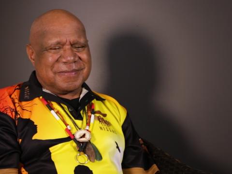 Archie Roach smiling, pictured from the waist up, seated and looking slightly away from camera.