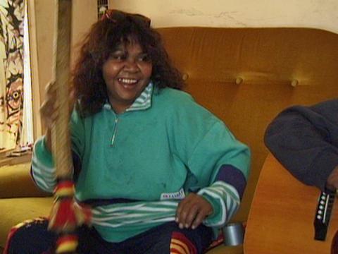 Ruby Hunter sitting on a sofa and smiling. She's holding an Aboriginal musical instrument.