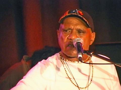 Archie Roach pictured from the chest up in front of a microphone, wearing a white t-shirt and black cap.