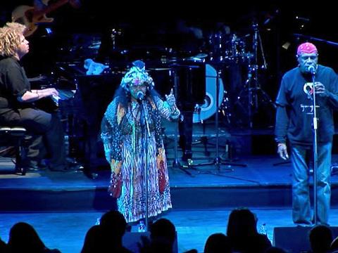 Indigenous musicians Archie Roach and Ruby Hunter pictured on stage,both are singing into microphones. There is a band in the background including a pianist, drummer and guitarist.