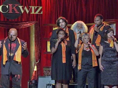 Indigenous singer-songwriter Archie Roach on stage performing for the SBS TV show Rockwiz. Also on stage are a choir of six people, including Aboriginal elder and performer, Uncle Jack Charles. 