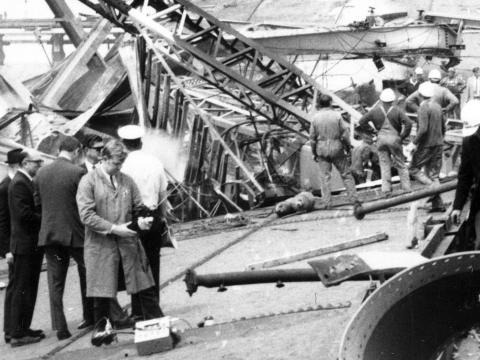 A black and white photo of the scene of the West Gate Bridge disaster. It shows investigators and bridge workers alongside the metal wreckage of the bridge.