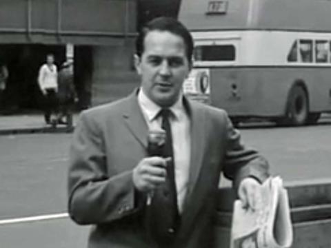 A TV reporter on a city street in the 1960s holding a microphone and a newspaper