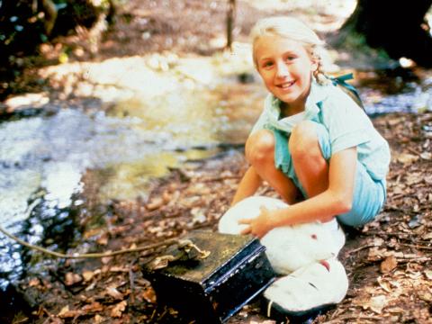 A young blonde-haired girl (Rebecca Smart) in blue shorts and shirt, crouching by the side of a stream with a white rabbit and a tradional japanese mask sitting at her feet.  