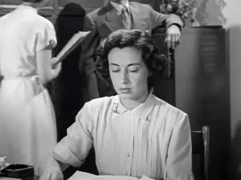 A woman is seated at an office desk reading aloud while behind her a man leans on a filing cabinet talking to another woman