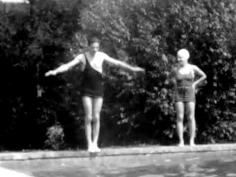 A man and woman in 1920s bathing suits standing by a swimming pool. The man has its arms out to his sides, about to jump in