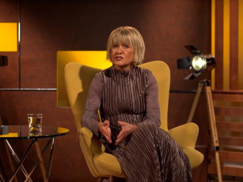 Still of Margaret Pomeranz talking to camera in a studio set up. There is a film light on a tripod and a glass of water on a table next to her.