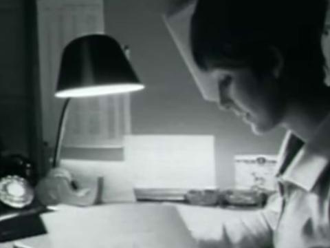 Black and white photo of a woman working at a desk. There is a black telephone and lamp on the desk.