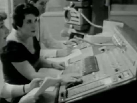 Two woman are sitting at a vision mixing desk in a television studio.