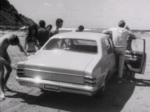 A group of young people stand around a Holden car on a beach