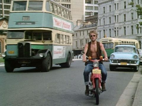 A man wearing a waistcoat and no shirt rides a motor bike down a city street. Two buses are behind him - one of which is a green and yellow double-decker