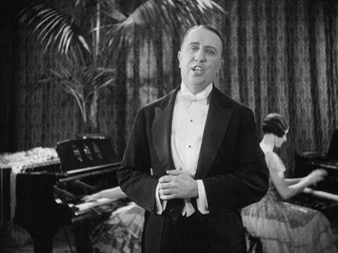 Singer Jack O'Hagan singing wearing a suit. a woman is playing the piano behind him.