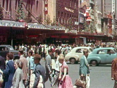 A crowd of people walking around a city street in Melbourne. Cars drive past in the background.