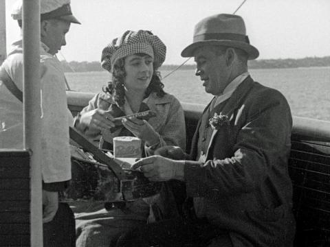 A scene from the film The Sentimental Block featuring Lottie Lyell and Arthur Tauchert sitting on a boat. A young boy stands in front of them as they talk to each other.
