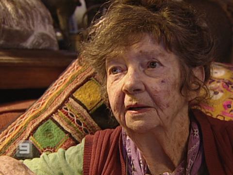 Artist Margaret Olley is sitting on a colourful couch being interviewed.