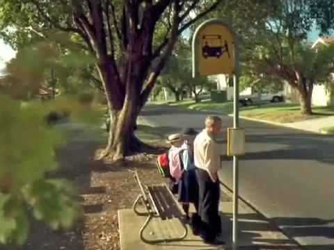 A man and his two children wait a bus stop in a leafy suburban street.