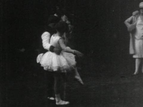 Black and white still of a girl dressed in ballet tutu and shoes. A boy has his arm around her waist and a woman looks on dressed in dress, hat and coat