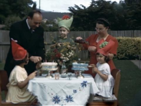 A family celebrate the birthdays of their two girls in the 1950s. Brother, mother and father watch on as the girls sit in front of their cakes.