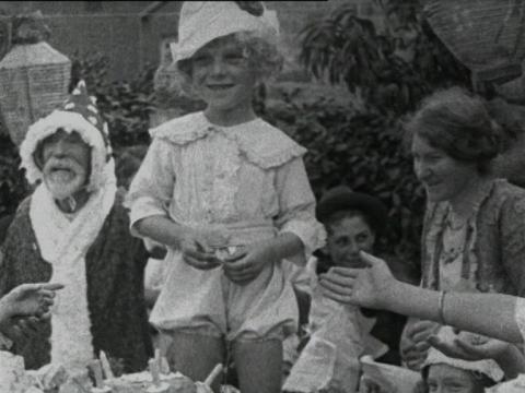 Black and white still of a man dressed as Santa and a young boy dressed in white, with a white hat on, standing beside a cake
