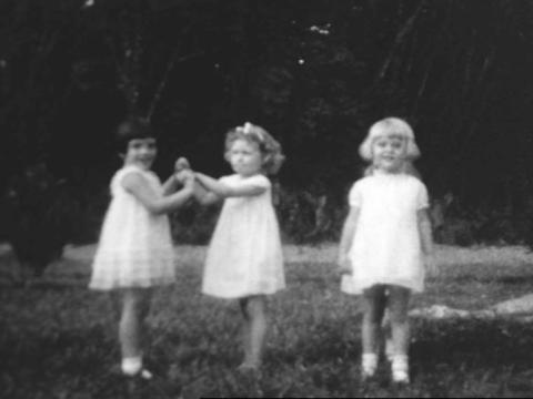Black and white still of three little girls wearing white dresses at a birthday party. Two of them are holding hands.