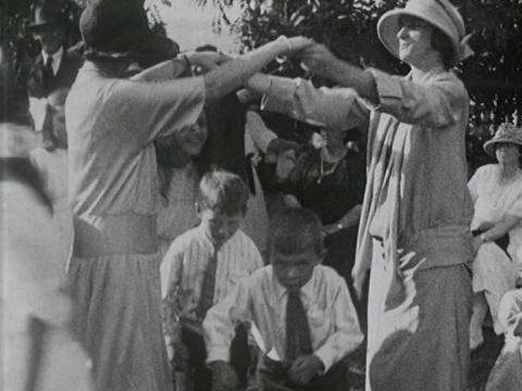 A black and white still depicting a children's party game called Oranges and Lemons where two women hold hands above the children who walk under them. They are dressed in 1920s clothing.