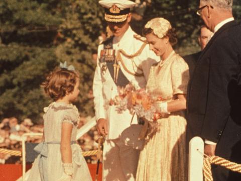 Queen Elizabeth II, with the Duke of Edinburgh standing to her right, receiving a bouquet of flowers from a young girl in 1954 during her first tour to Australia.