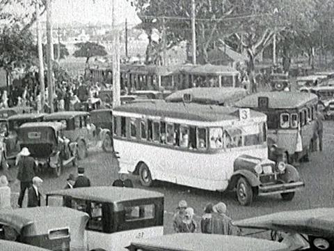 Anzac Parade Junction, circa 1928, with crowds of people walking among cars, buses and trams.