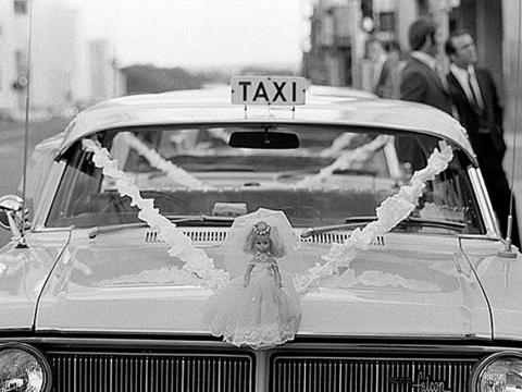 Black and white image of front of a taxi with a doll in a wedding dress sitting on the hood of the car.