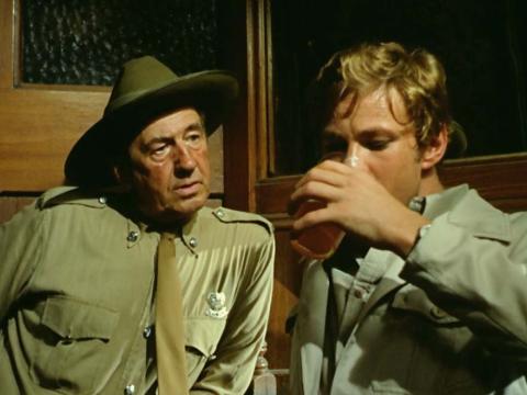 John Grant, played by Gary Bond, drinks a beer as policeman Jock Crawford , played by Chips Rafferty, looks on.