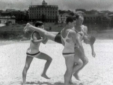 Life guards at Bondi Beach performing a rescue demonstration. Three of the life guards carry a man along the beach by his legs and torso.  