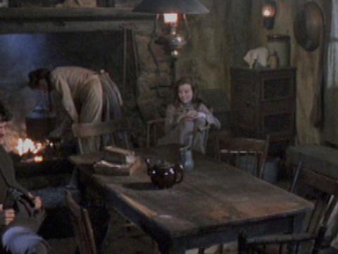 The Kelly family in the kitchen of their farmhouse.. Some stand, some sit at the table. Mother tends the fire.