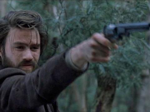 Heath Ledger as Ned Kelly holds a gun out in front of him.