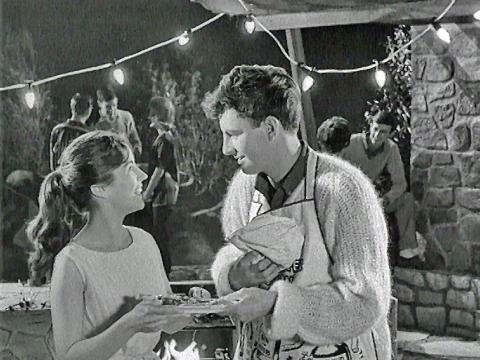 A young woman and young man standing in an outdoor setting in front of a barbecue holding a plate of food. There are other young people socialising in the background.