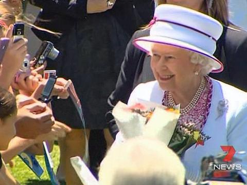 Queen Elizabeth II wearing a white hat and jacket, meeting and accepting flowers from a crowd of people.