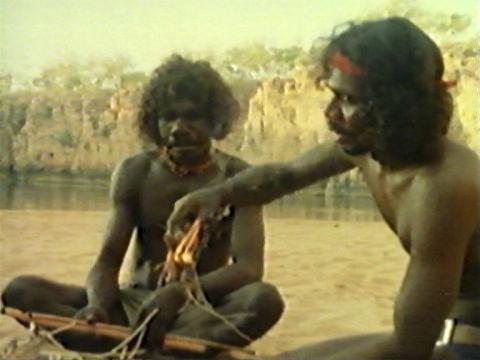 Actor David Gulpilil and another Aboriginal man sitting on the ground in the outback. Gulpilil is holding some kindling which is alight.