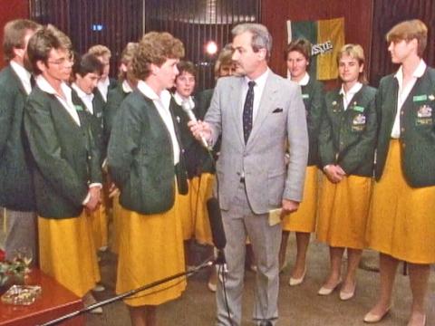 The 1987 Women's test cricket team standing in a semi-circle, wearing yellow skirts and green blazers. One of the women is being interviewed by a news reporter holding a microphone.