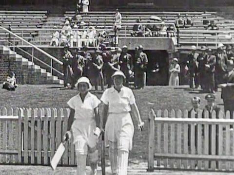 Two women cricketers walking onto a cricket pitch, they are wearing white uniforms and protective leg pads and holding cricket bats.