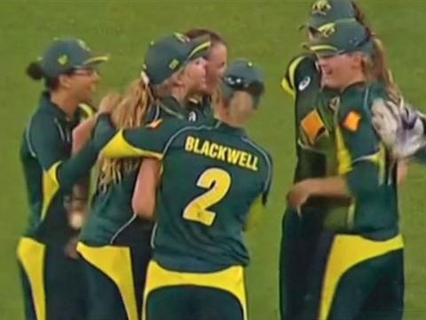 A group of Australian women cricketers gather standing around in a group on the cricket field in their uniforms smiling and hugging eachother.