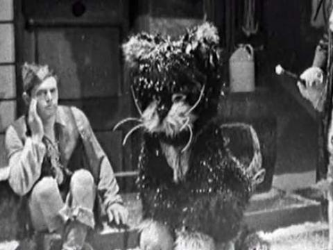 A young man in period costume sits on a doorstep next to a person in a large cat costume