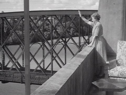 A woman waves to someone from a balcony. A suspension bridge is directly beside her.