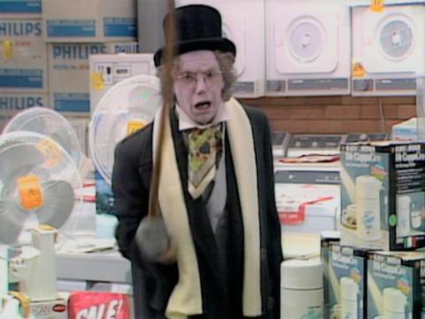A man in bad make-up that is meant to make him look older shaking a walking stick at the camera. He has a lot of home appliances behind him.