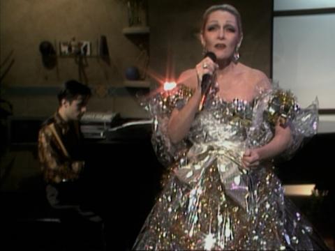 Jeanne Little wearing an elaborate silver and gold dress. She is singing into a microphone. John Foreman is in the background playing piano.