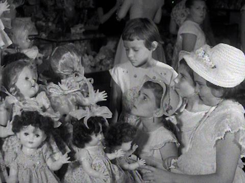 A little girl looks up at a range of toy dolls with pleading eyes in a black and white still. She is surrounded by three other girls in white dresses.