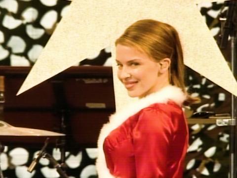 Kylie Minogue looks over her shoulder cheekily at the audience. She is on a stage. She is dressed in a Christmas outfit with red silk and fur trim.
