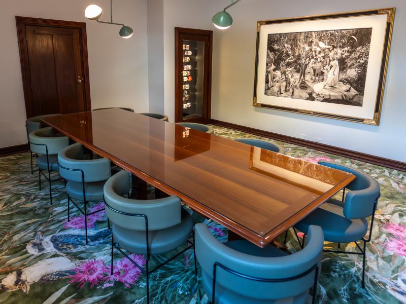 A meeting room with a colourful carpet and blue chairs arranged around a long wooden boardroom table. There is a framed photograph on the wall.