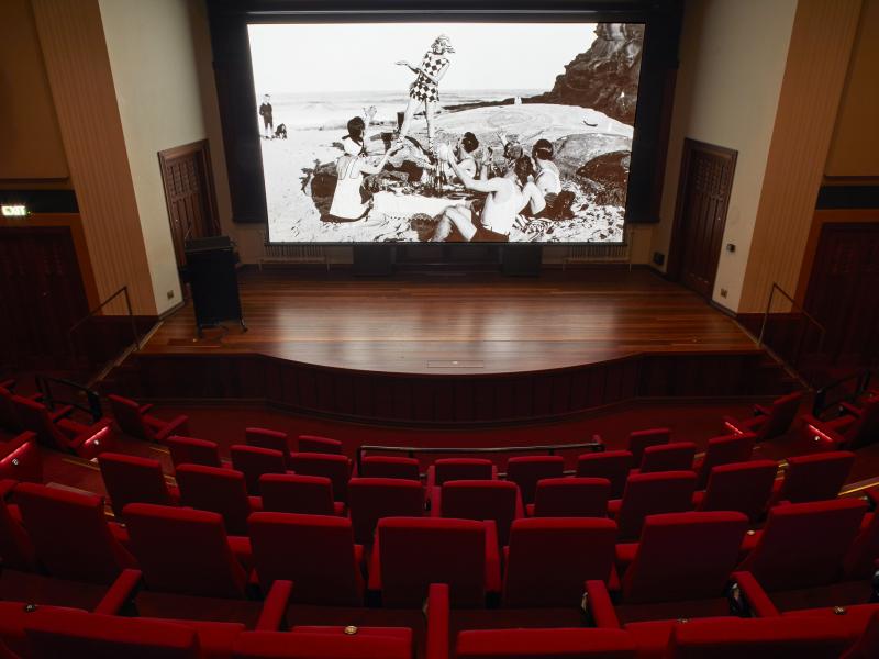 View from the back row of an empty cinema with red seating, large screen and wooden stage at front 