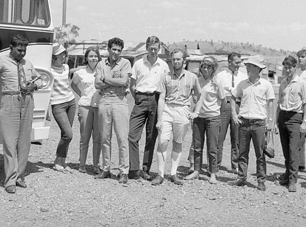 A group of 13 people standing side by side next to the front of a bus looking towards the camera.