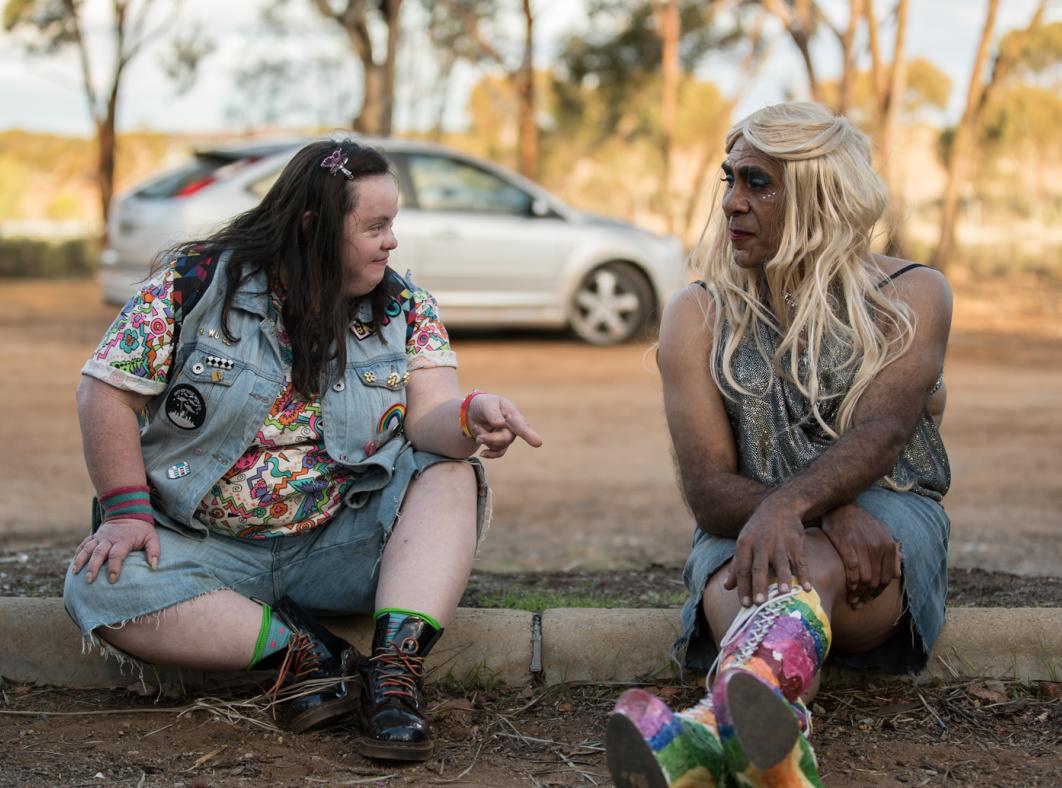 A young woman with Down Syndrome sits beside a drag queen on the ground in front of a parking lot.