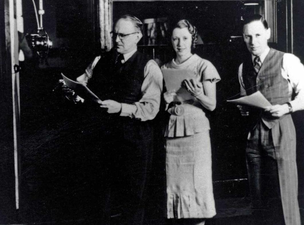 Two men and a woman in a recording studio standing next to a large microphone.