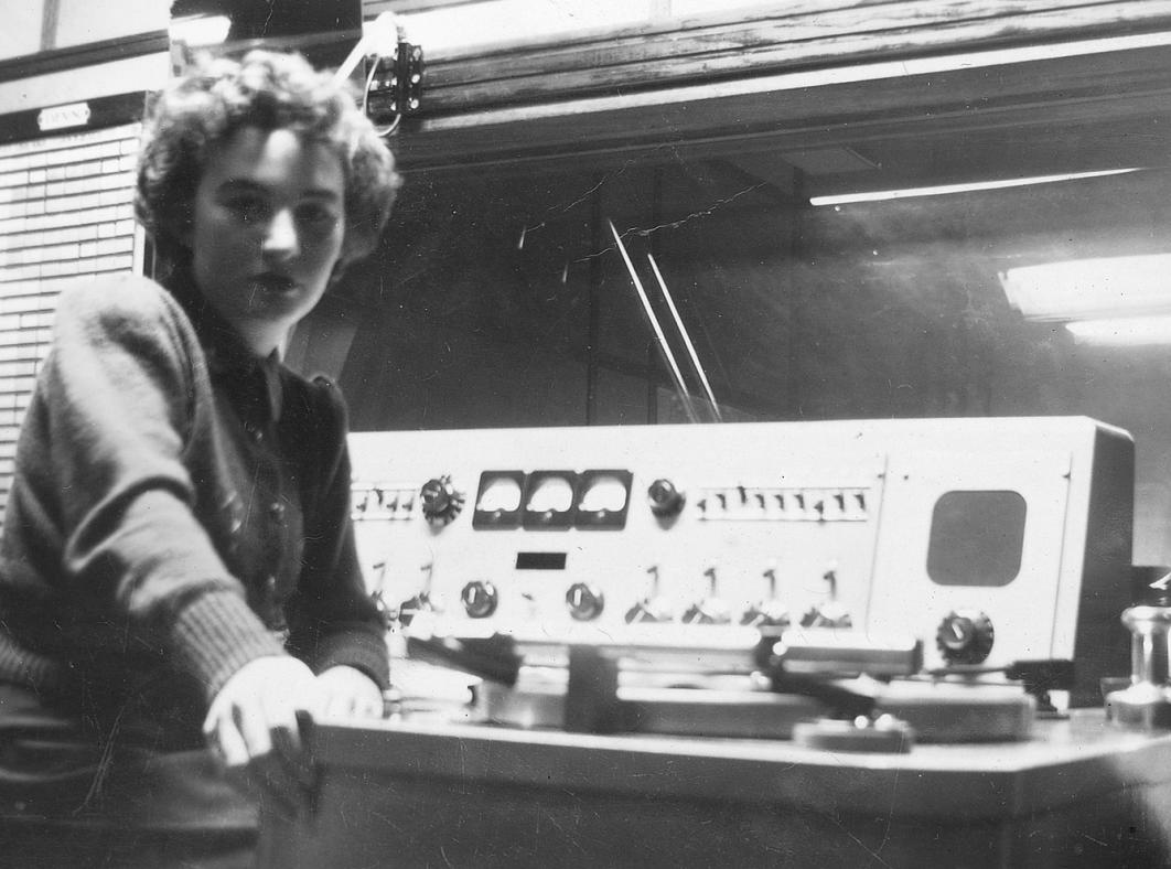 A woman in a radio studio with sound equipment beside her, she is turned sideways to face the camera.